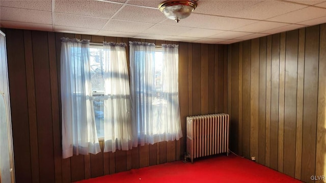 carpeted spare room with radiator, a drop ceiling, and wood walls