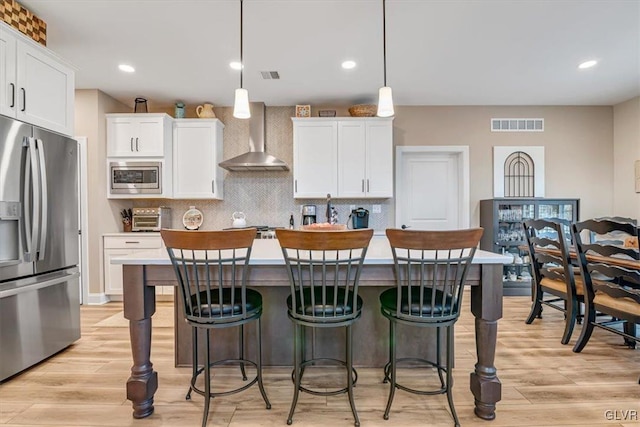 kitchen with appliances with stainless steel finishes, pendant lighting, white cabinetry, an island with sink, and wall chimney exhaust hood