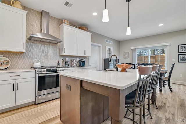 kitchen featuring stainless steel gas range, white cabinetry, a center island with sink, pendant lighting, and wall chimney range hood