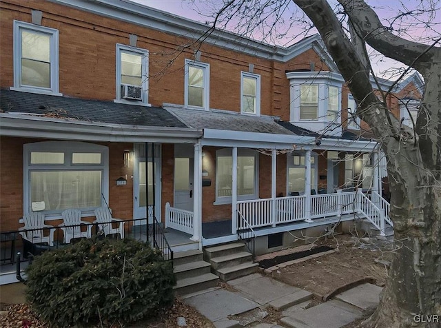 townhome / multi-family property with a shingled roof, a porch, cooling unit, and brick siding