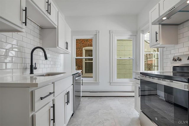 kitchen with baseboard heating, appliances with stainless steel finishes, white cabinetry, a sink, and wall chimney exhaust hood