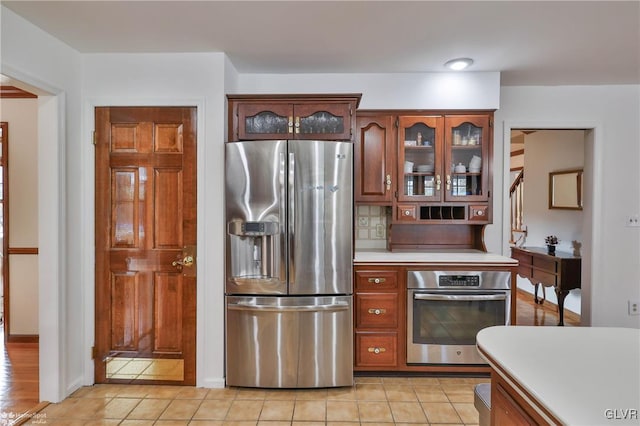 kitchen with appliances with stainless steel finishes, light countertops, glass insert cabinets, and tasteful backsplash