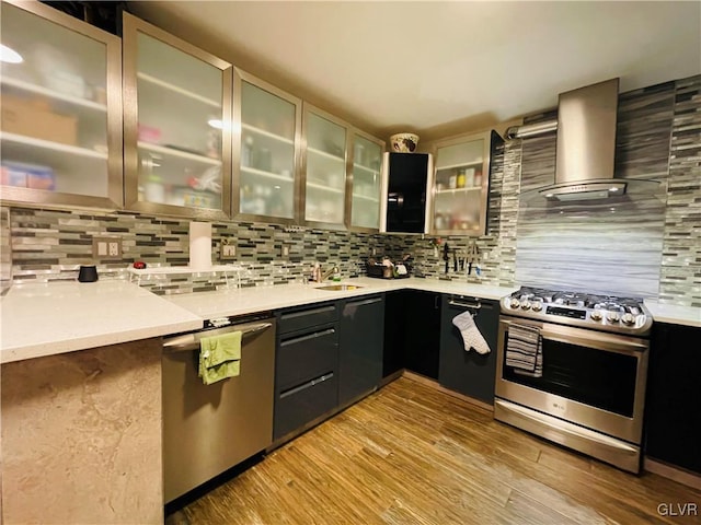 kitchen with light countertops, appliances with stainless steel finishes, wall chimney range hood, backsplash, and glass insert cabinets