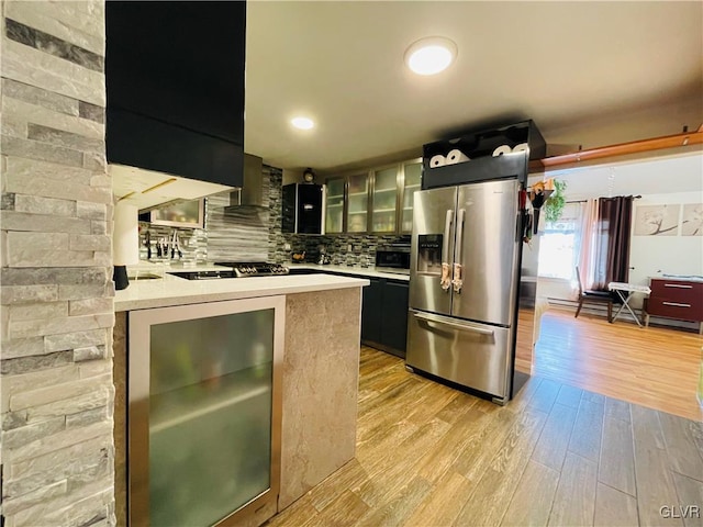 kitchen featuring light wood finished floors, stainless steel fridge with ice dispenser, glass insert cabinets, light countertops, and wall chimney range hood