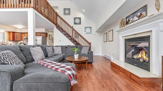 living room with stairway, a baseboard heating unit, a glass covered fireplace, wood finished floors, and high vaulted ceiling