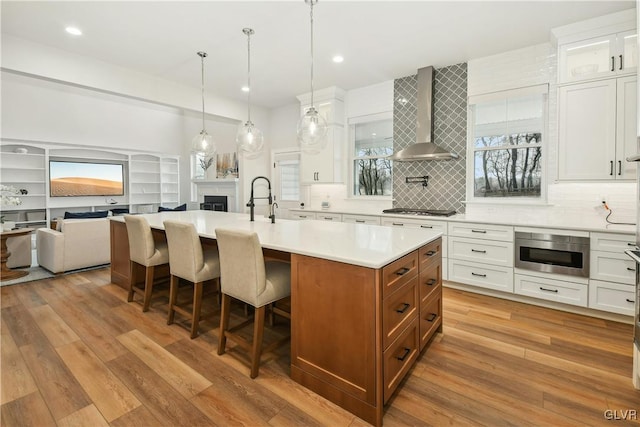 kitchen featuring an island with sink, wall chimney exhaust hood, open floor plan, light countertops, and white cabinetry