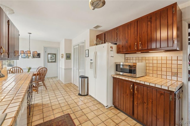 kitchen with white refrigerator with ice dispenser, decorative backsplash, tile countertops, stainless steel microwave, and decorative light fixtures