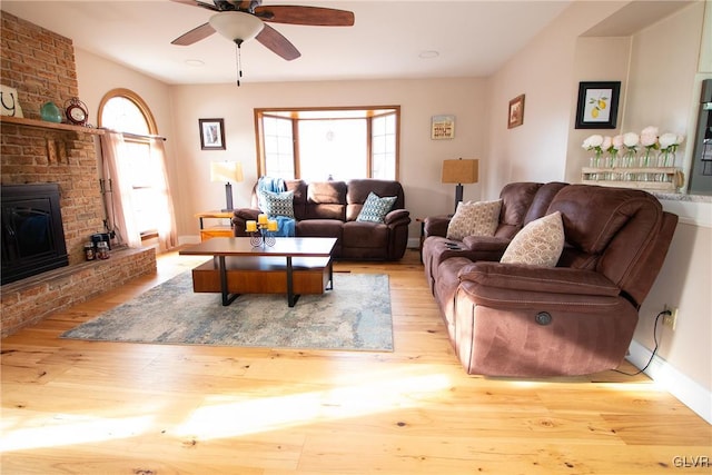 living area featuring a brick fireplace, light wood-style flooring, baseboards, and ceiling fan