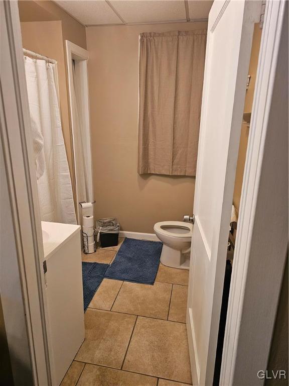 bathroom with a drop ceiling, tile patterned flooring, toilet, vanity, and baseboards