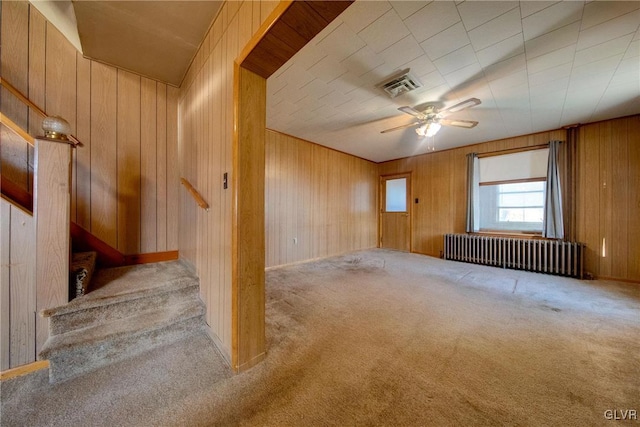 carpeted empty room with visible vents, radiator heating unit, ceiling fan, stairway, and wood walls