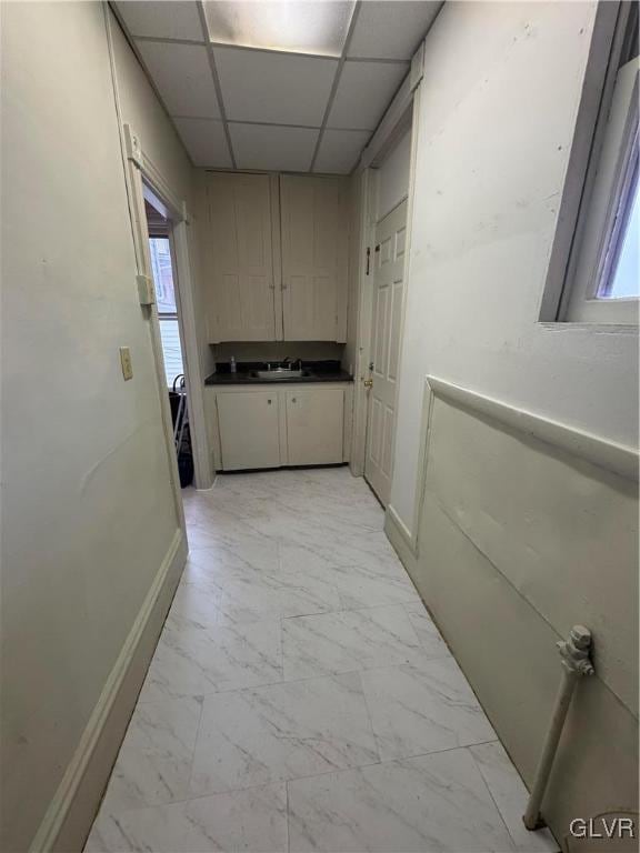 hall with a paneled ceiling, marble finish floor, baseboards, and a sink