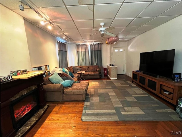 living room featuring rail lighting, a ceiling fan, a drop ceiling, a lit fireplace, and baseboards