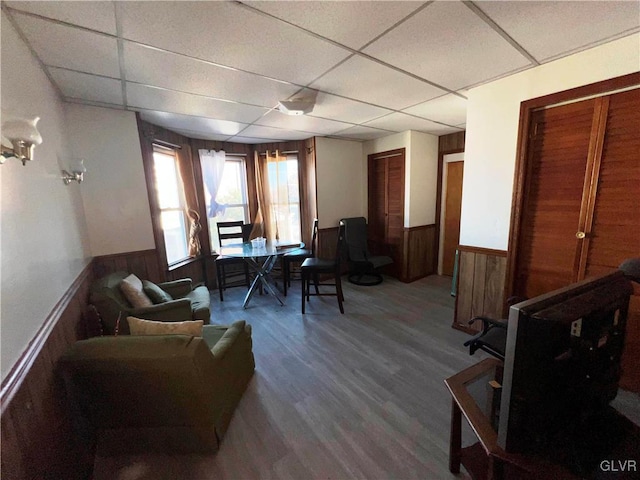 living area with a wainscoted wall, wood walls, a drop ceiling, and wood finished floors