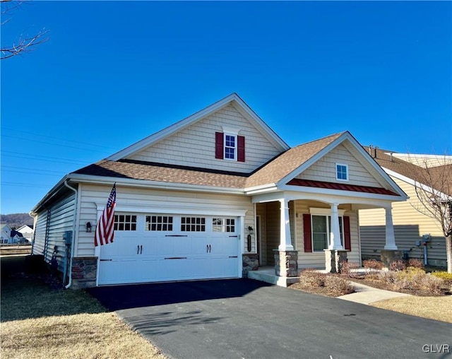 craftsman-style home featuring aphalt driveway, covered porch, a garage, a shingled roof, and stone siding