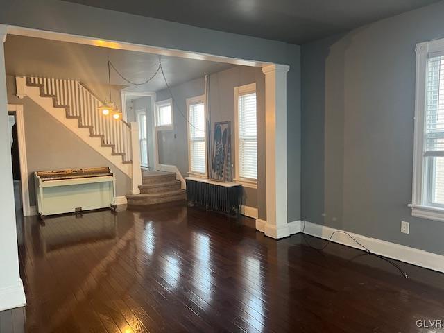 unfurnished living room featuring baseboards, stairway, radiator, wood-type flooring, and ornate columns