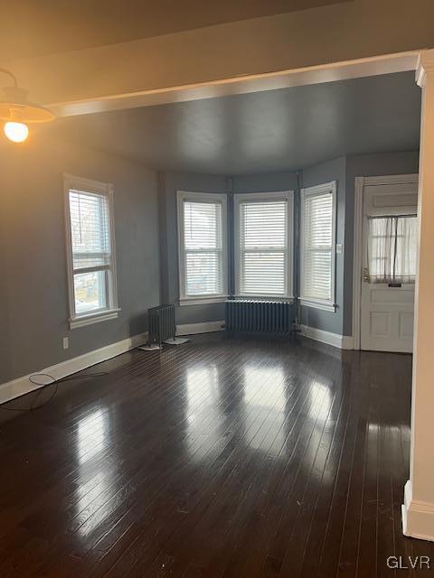 unfurnished living room with dark wood-type flooring, a healthy amount of sunlight, baseboards, and radiator heating unit