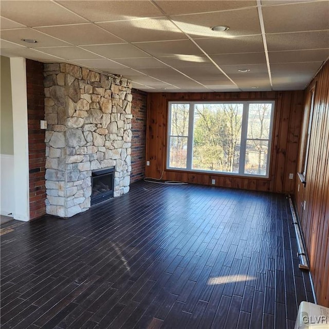 unfurnished living room featuring dark wood-style floors, a drop ceiling, wood walls, and a stone fireplace