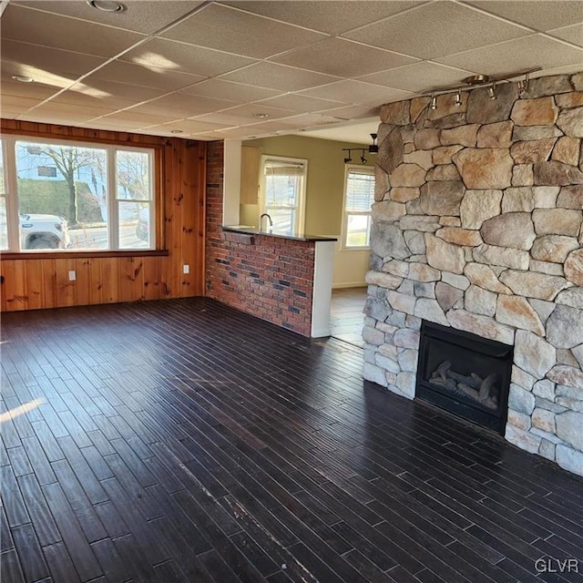 unfurnished living room featuring dark wood-style floors, a stone fireplace, a drop ceiling, and wooden walls