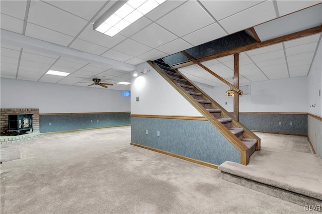 finished basement with a drop ceiling, a wainscoted wall, a wood stove, stairs, and carpet floors