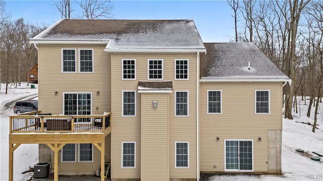 snow covered back of property featuring cooling unit, roof with shingles, and a wooden deck