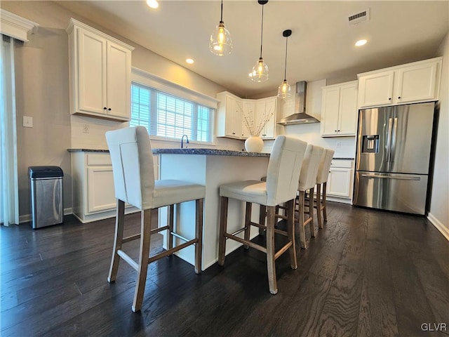 kitchen with dark wood-style flooring, stainless steel refrigerator with ice dispenser, visible vents, white cabinetry, and wall chimney exhaust hood