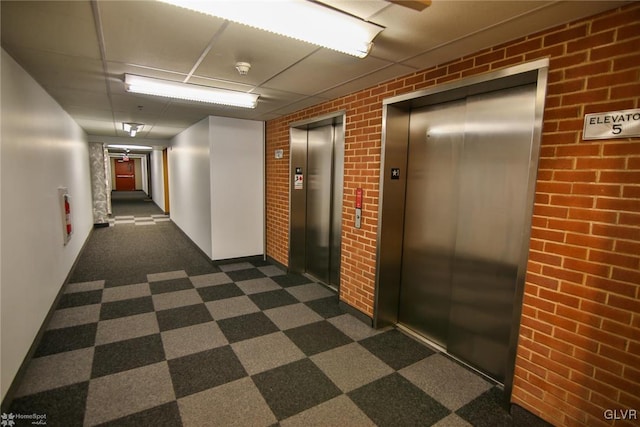 hall with elevator, a drop ceiling, and brick wall