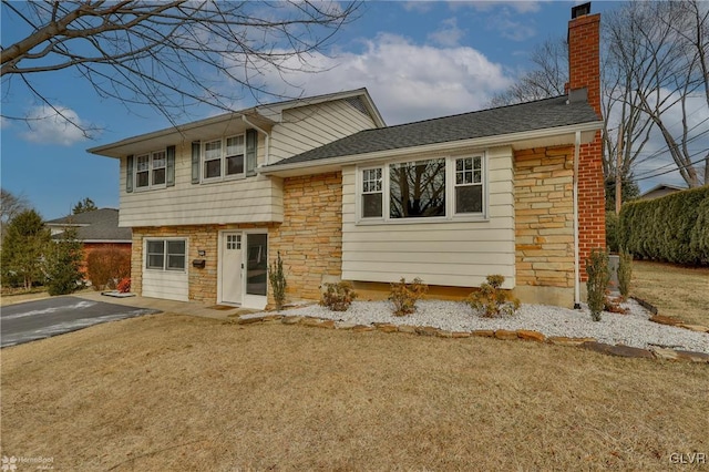 tri-level home with a shingled roof, stone siding, a chimney, aphalt driveway, and a front yard