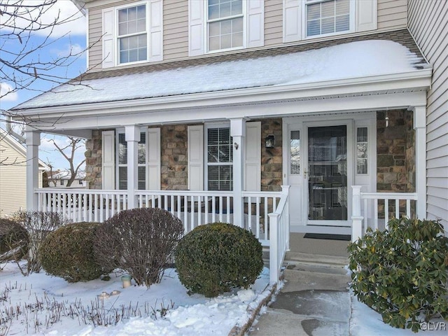 snow covered property entrance featuring stone siding, covered porch, and roof with shingles