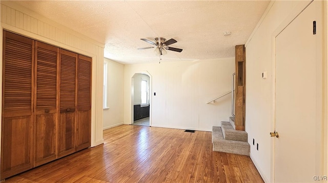 unfurnished room with a textured ceiling, wood finished floors, arched walkways, ceiling fan, and stairs