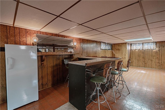 bar featuring tile patterned floors, freestanding refrigerator, a bar, wood walls, and a paneled ceiling