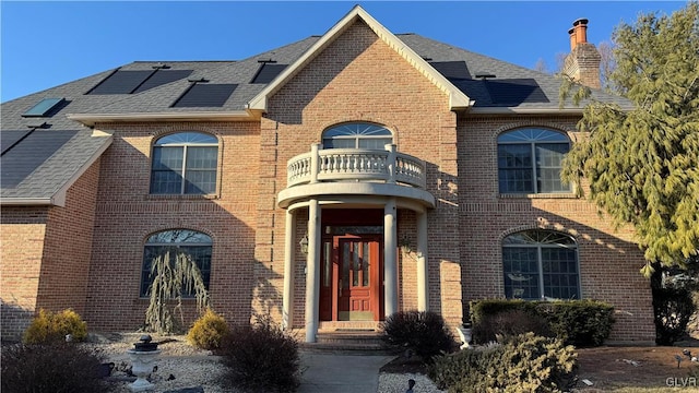 view of front facade with brick siding, a shingled roof, roof mounted solar panels, a chimney, and a balcony