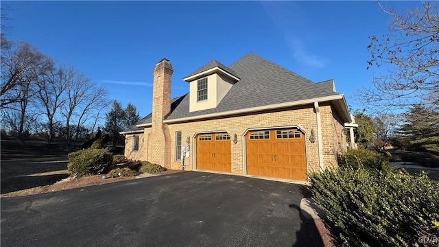 view of side of home featuring driveway, a shingled roof, a garage, brick siding, and a chimney