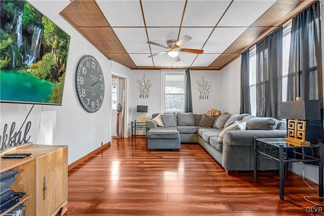 living area with ceiling fan, wood finished floors, baseboards, and a paneled ceiling