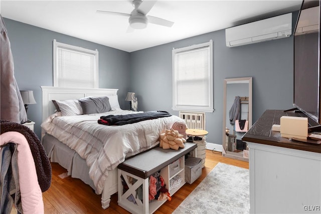 bedroom featuring a wall mounted AC, wood finished floors, radiator heating unit, baseboards, and ceiling fan