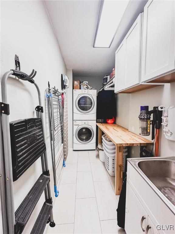 clothes washing area featuring light tile patterned flooring, cabinet space, stacked washing maching and dryer, and a sink