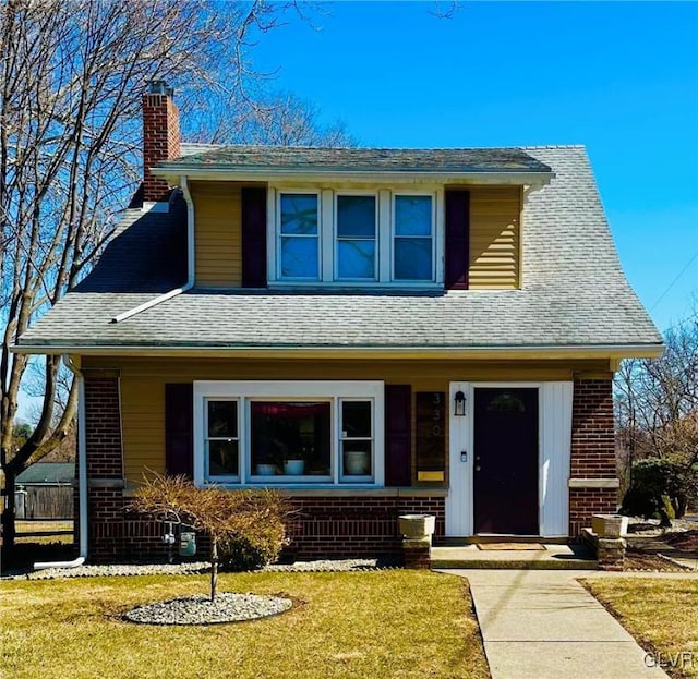 view of front of home with a front yard, brick siding, roof with shingles, and a chimney