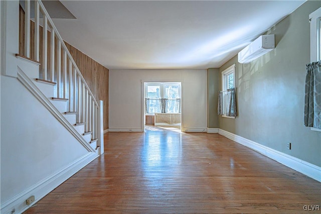 unfurnished living room with stairs, a wall mounted AC, baseboards, and wood finished floors