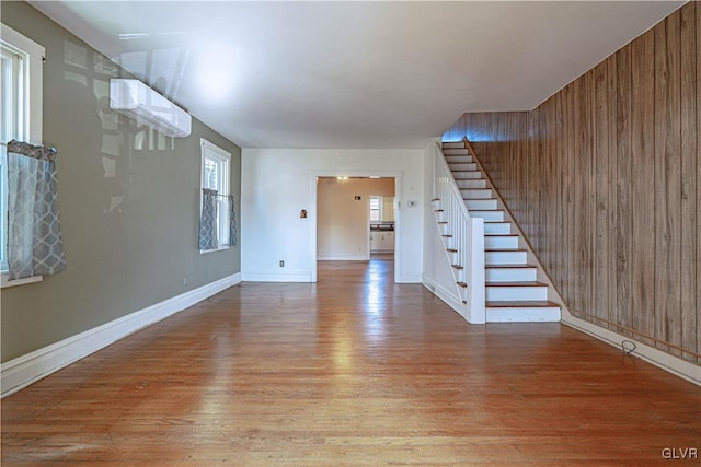 unfurnished living room with wooden walls, baseboards, stairs, a wall unit AC, and wood finished floors