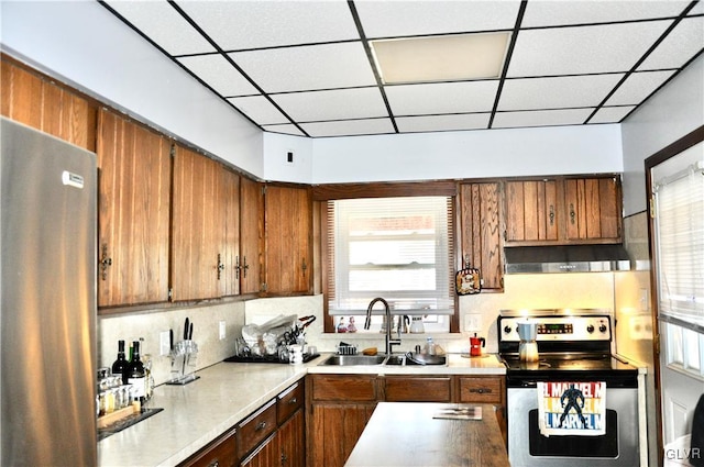 kitchen with a sink, light countertops, appliances with stainless steel finishes, wall chimney range hood, and brown cabinets