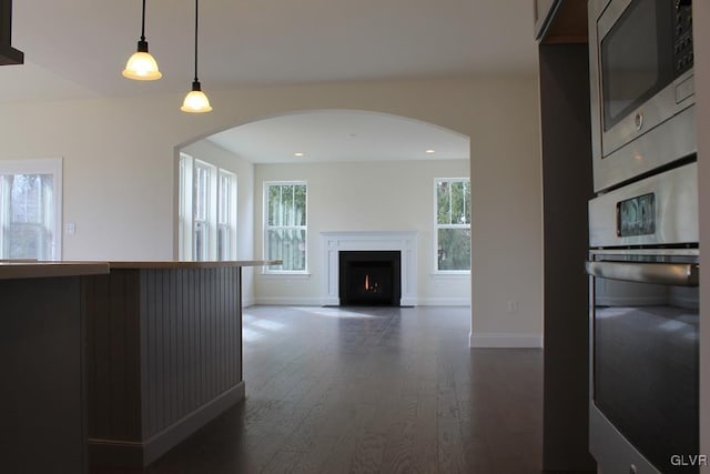 kitchen featuring baseboards, dark wood finished floors, a lit fireplace, arched walkways, and stainless steel appliances