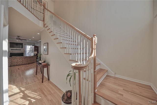 staircase with ceiling fan, baseboards, a fireplace, a high ceiling, and wood finished floors