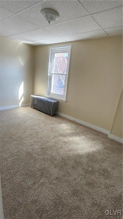 empty room with carpet flooring, radiator heating unit, baseboards, and a drop ceiling