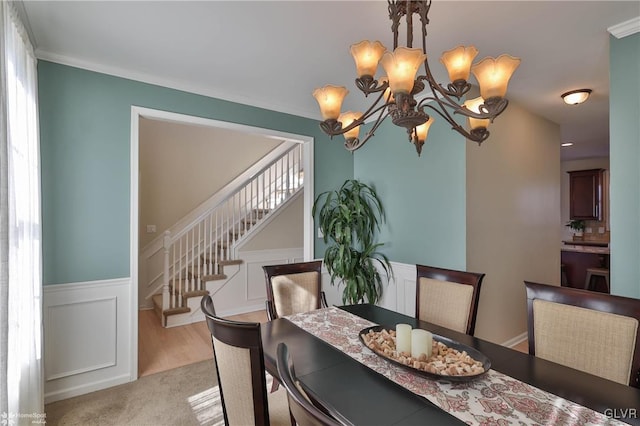 dining room featuring a wainscoted wall, stairs, a decorative wall, light colored carpet, and a chandelier