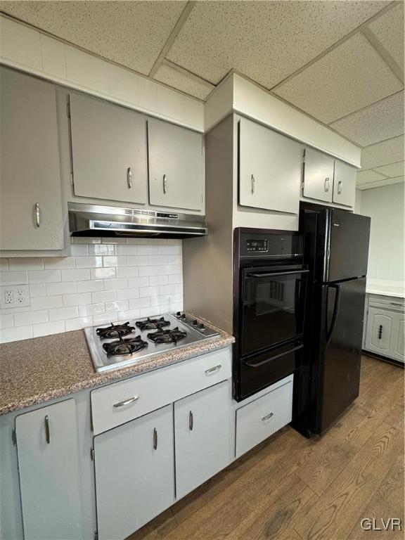 kitchen featuring a warming drawer, black appliances, under cabinet range hood, tasteful backsplash, and dark wood-style flooring