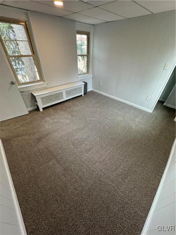 empty room with a drop ceiling, wooden walls, radiator, carpet flooring, and baseboards