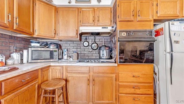 kitchen featuring brown cabinetry, light countertops, under cabinet range hood, appliances with stainless steel finishes, and tasteful backsplash