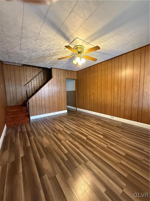 unfurnished living room featuring dark wood-style floors, baseboards, ceiling fan, stairs, and wood walls