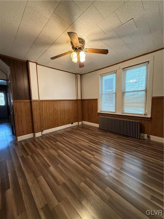 empty room with dark wood-style floors, wainscoting, radiator heating unit, and a ceiling fan