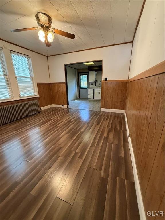 empty room with a ceiling fan, wood finished floors, wood walls, wainscoting, and crown molding