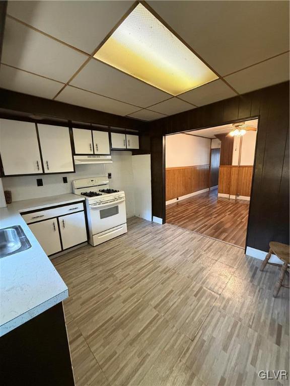 kitchen with wooden walls, white range with gas stovetop, light countertops, a paneled ceiling, and under cabinet range hood